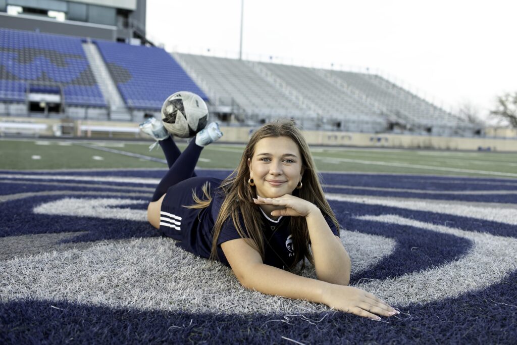 High school soccer player resting a foot on the ball with the bleachers behind her, golden hour lighting glowing.