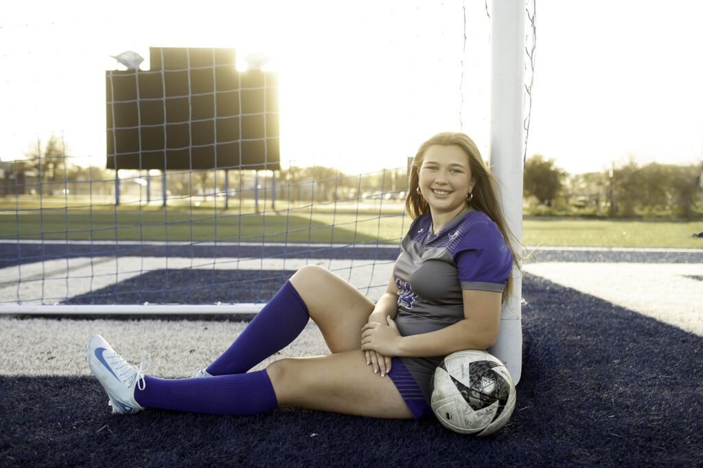 ​High school athlete sitting on the ground near the goal, leaning back on her hands, with a soccer ball resting beside her and the netting behind.