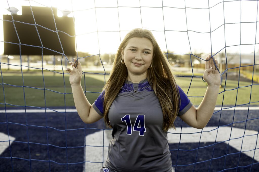 Teen soccer player holding onto the goal net, looking up with a thoughtful expression, the goal netting softly blurred around her.