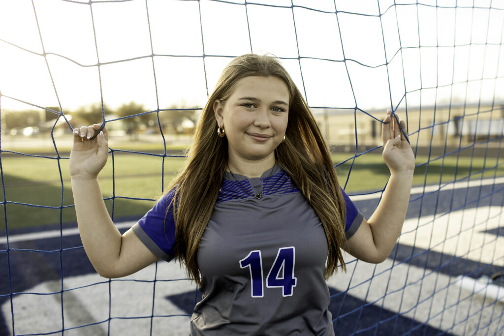 Teen soccer athlete holding onto the goal net, leaning forward slightly, with the netting framing her face and the field blurred behind her.