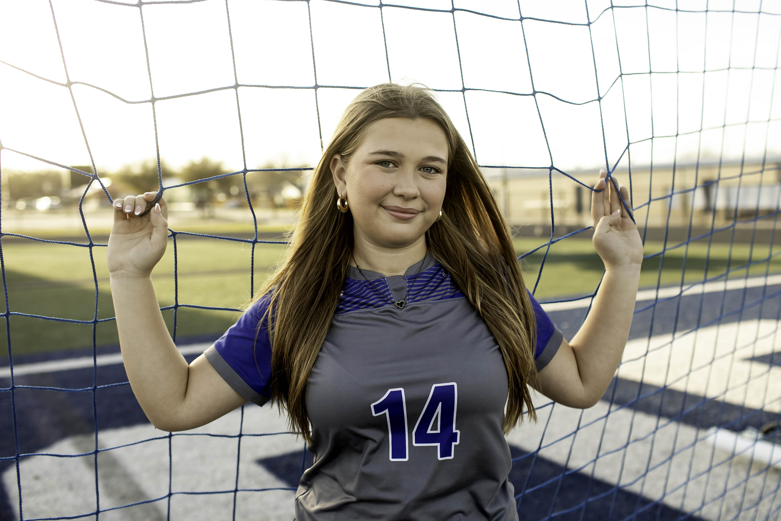 Teen soccer athlete holding onto the goal net, leaning forward slightly, with the netting framing her face and the field blurred behind her.