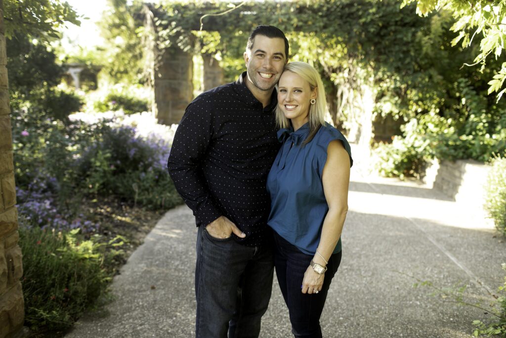 Parents standing close together, holding hands and smiling at each other with the gardens in the background