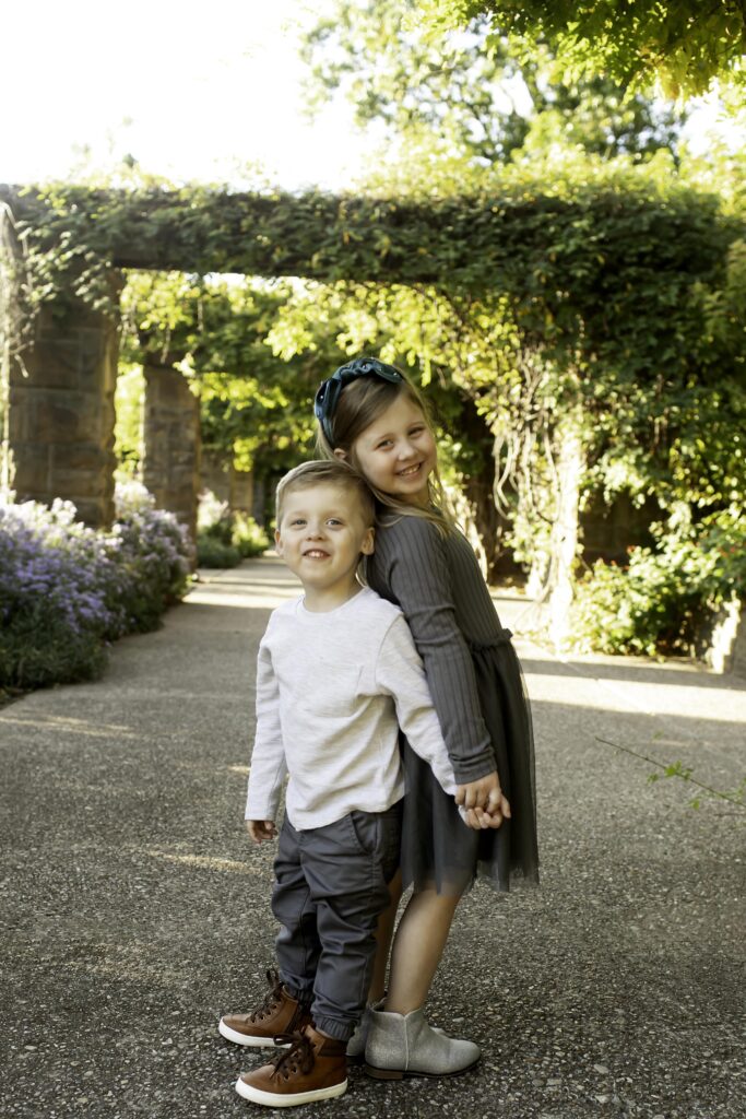 Brother and sister standing back-to-back, looking over their shoulders with playful grins