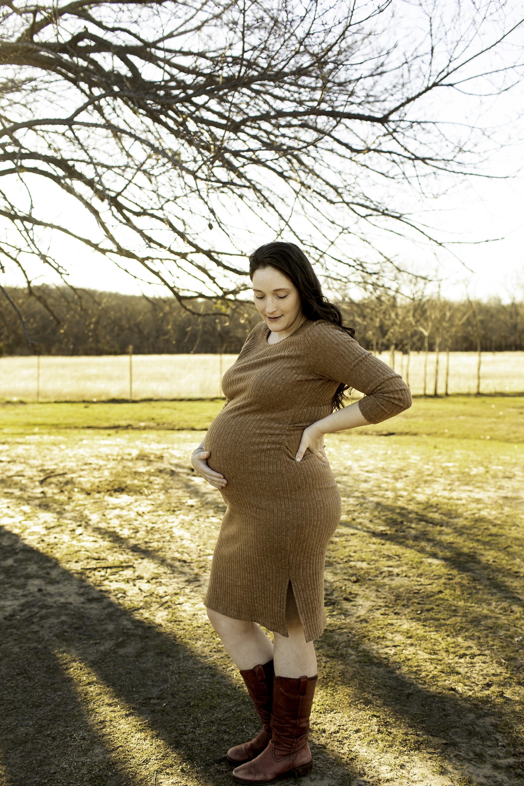 Expecting mom standing in the middle of a farm field, wearing a dress, her hands resting gently on her belly as the sun sets behind her.