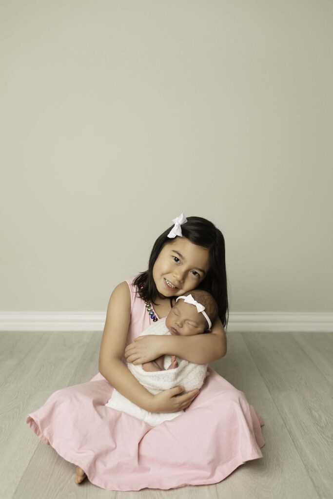 Big sister gently cradling her newborn sister, both wrapped in soft pastel blankets