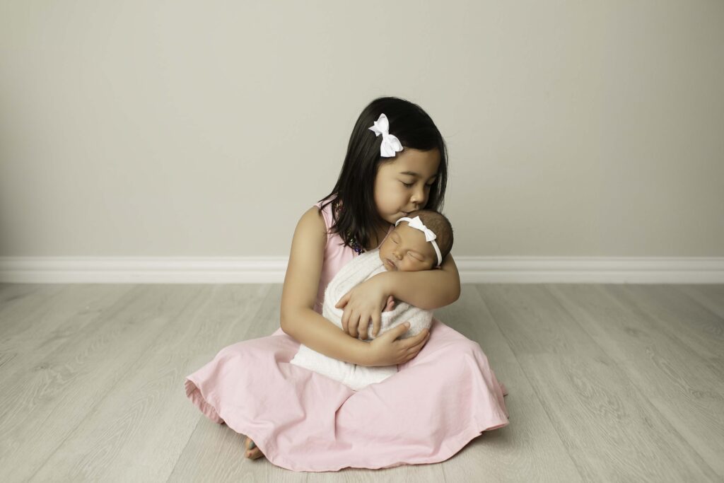 Big sister kissing her newborn sister's forehead, both snuggled together in natural light.