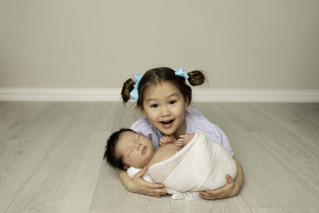 Big sister snuggling her newborn baby brother, both looking content and peaceful together.