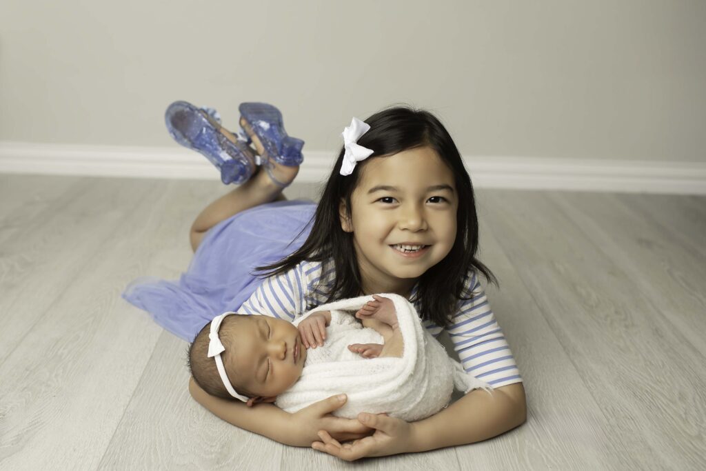 Big sister smiling at the camera while cuddling her newborn sister in her arms