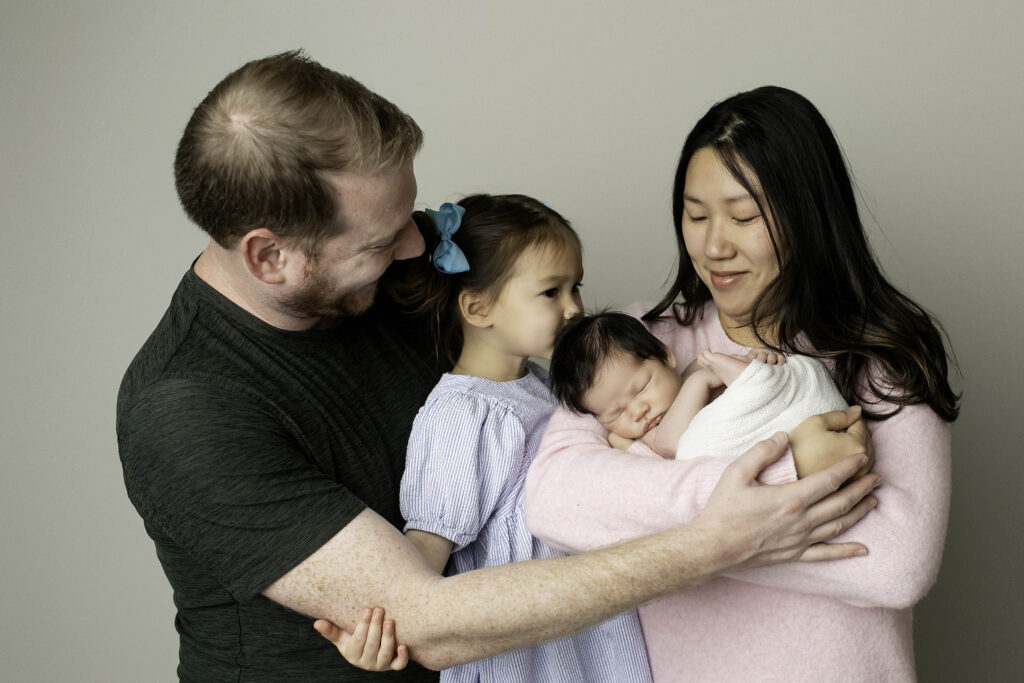 Studio portrait of dad, mom, big sister, and newborn brother, with the family gathered closely together, parents holding the baby while the sister smiles beside them