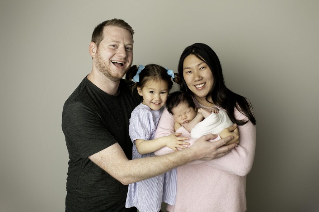 Family of four in the studio: dad and mom sitting together, with big sister standing next to them while holding their newborn baby brother, all smiling lovingly